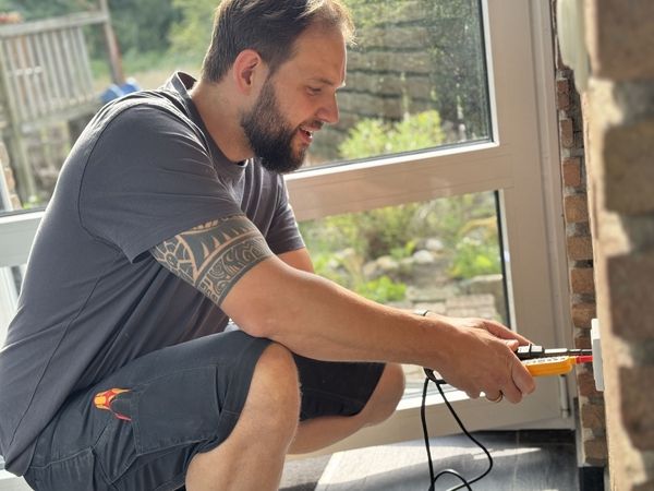 Mann mit tätowiertem Arm kniet neben einer Wand und hält ein Messgerät in der Hand. Er trägt ein graues T-Shirt und schwarze Shorts.