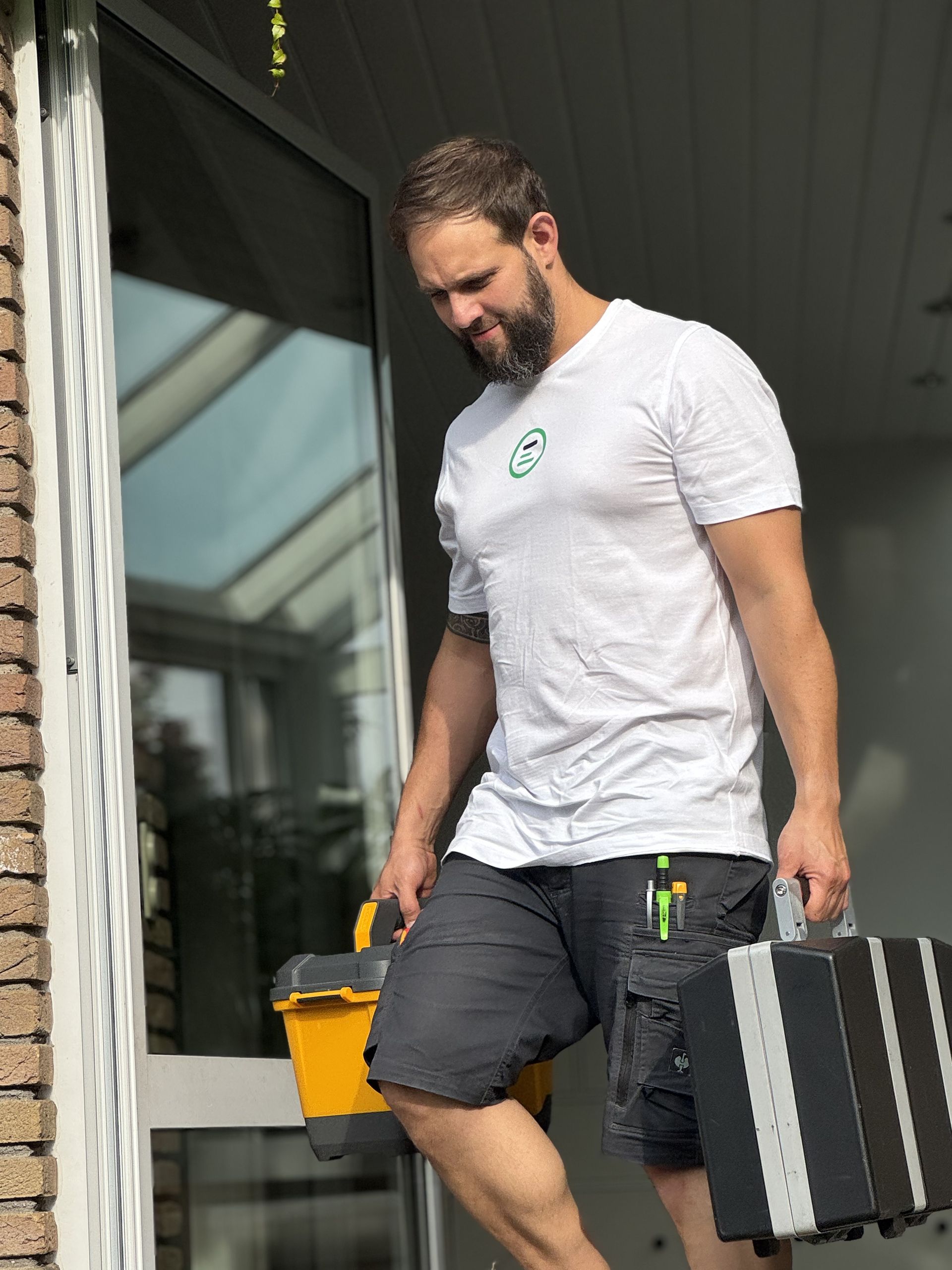 Mann in weißem T-Shirt und schwarzer Shorts trägt gelbe Werkzeugkiste und schwarzen Werkzeugkoffer mit silbernen Streifen