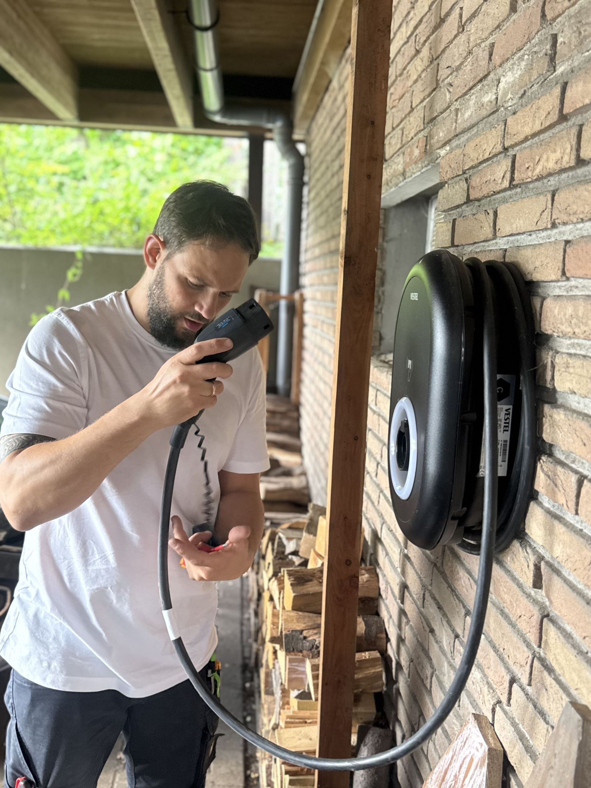 Ein Mann steht vor einer Wand mit einer schwarzen Ladestation für Elektrofahrzeuge. Er hält ein Kabel in der Hand und schaut auf ein Gerät.