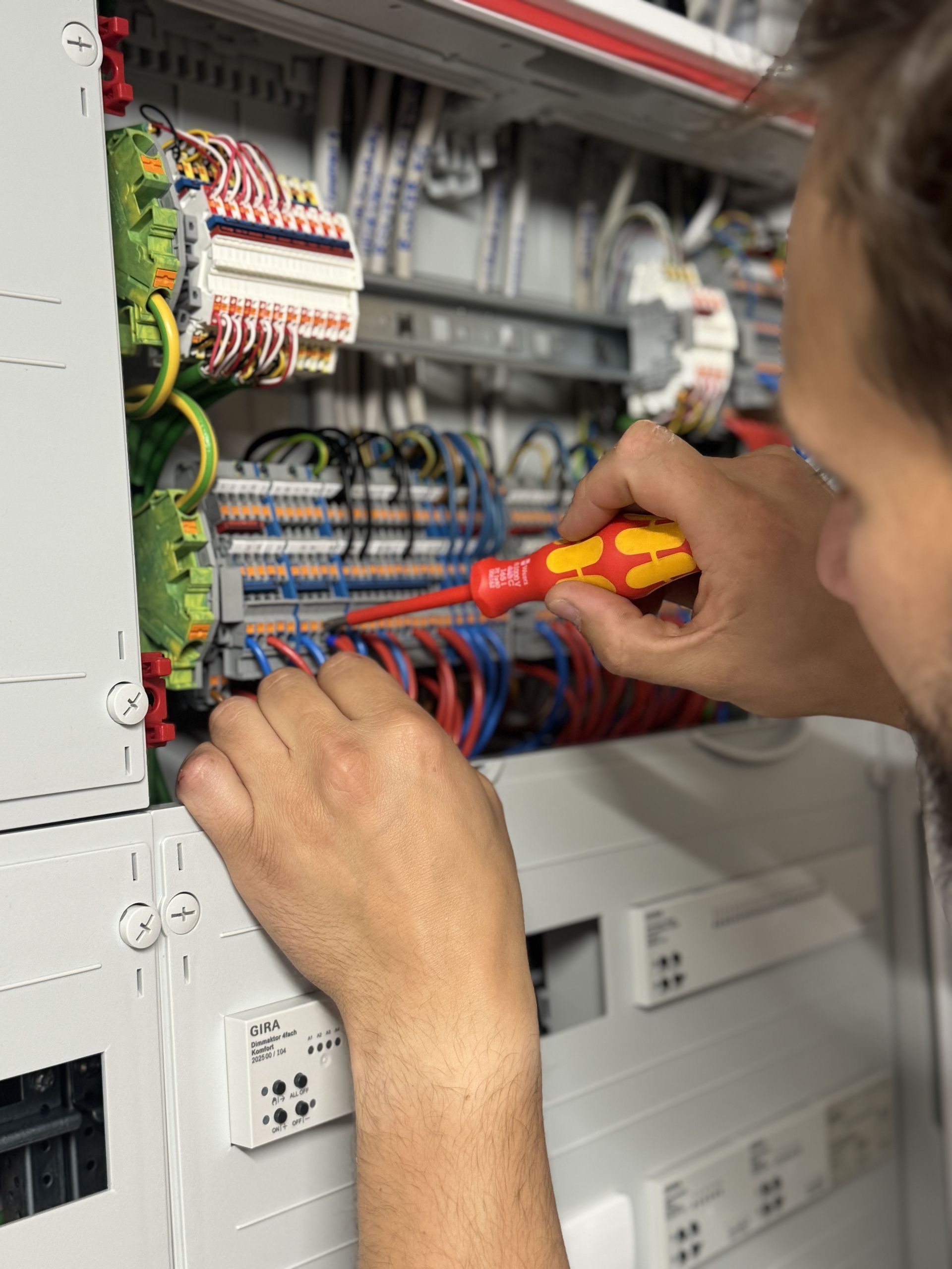 Hand hält einen Schraubendreher und arbeitet an einem elektrischen Schaltschrank mit vielen Kabeln und Sicherungen
