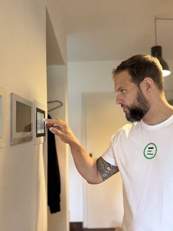 Ein Mann mit einem weißen T-Shirt und einem Tattoo am Arm berührt einen digitalen Bildschirm an der Wand. Im Hintergrund ist ein schwarzer Kleiderhaken sichtbar.