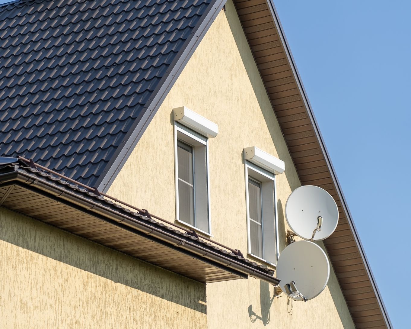 Dach eines Einfamilienhauses mit dunkler Dacheindeckung, Schornstein und zwei Satellitenschüsseln an der Fassade.